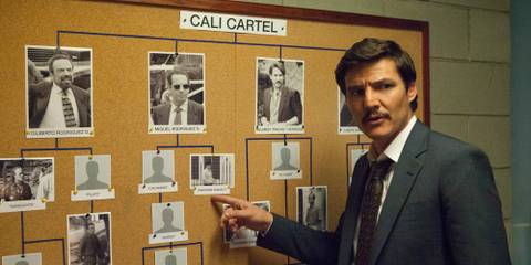 Pedro Pascal as Pablo Escobar pointing at a board full of pictures, while looking over his shoulder and talking to someone, in Narcos.