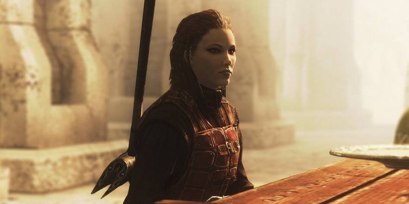 Ingjard sitting at a table in Skyrim, wearing leather armor and wielding a large weapon on her back.