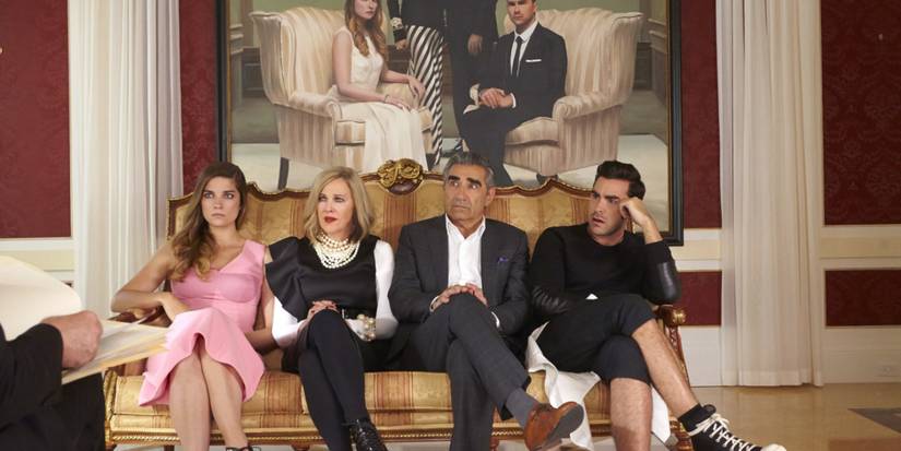 The Rose Family sit on a fancy sofa in front of a family portrait in the pilot episode of Schitt's Creek.