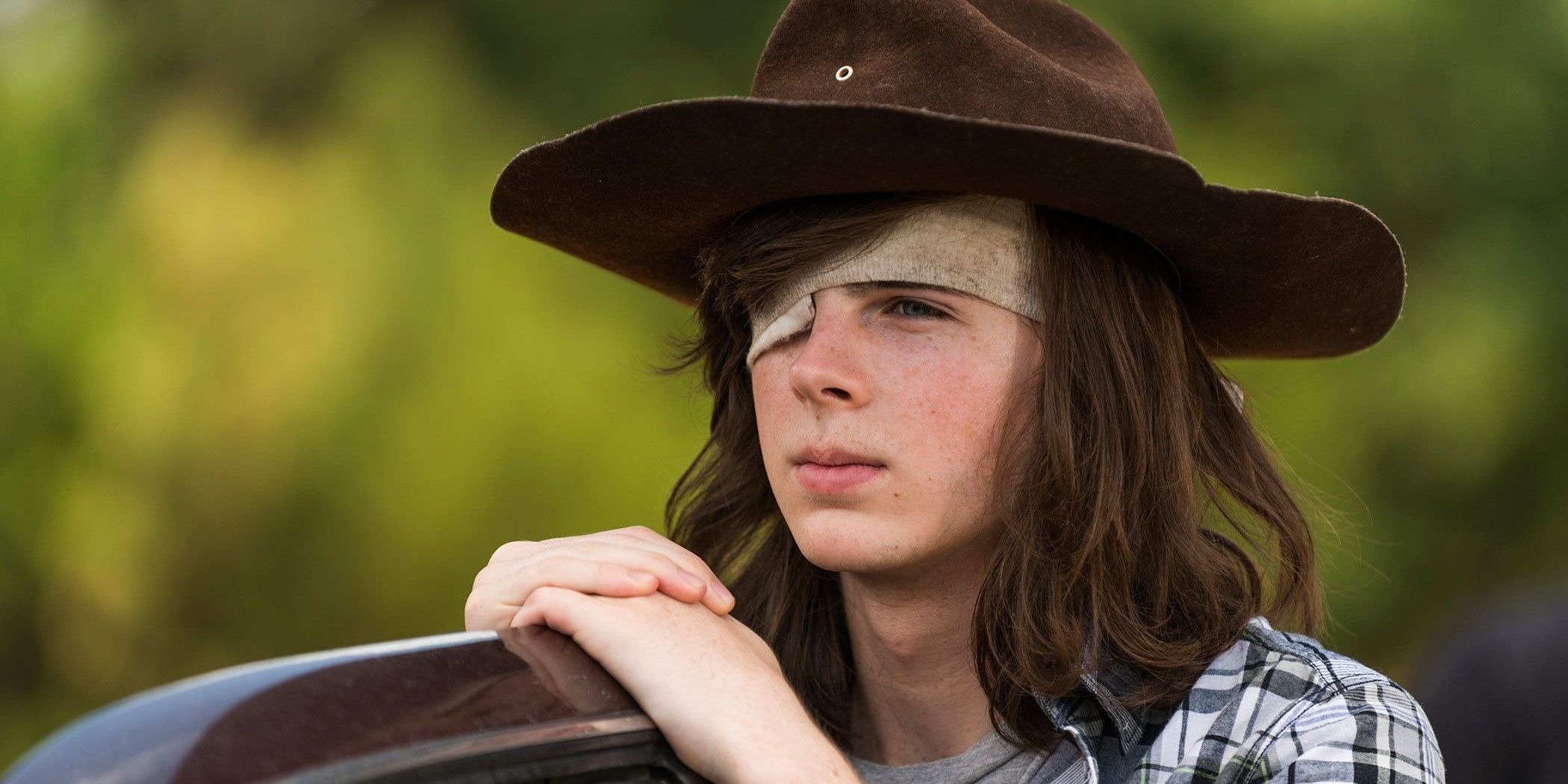 Carl Grimes (Chandler Riggs)wearing a hat and eye-patch while leaning on an object in The Walking Dead