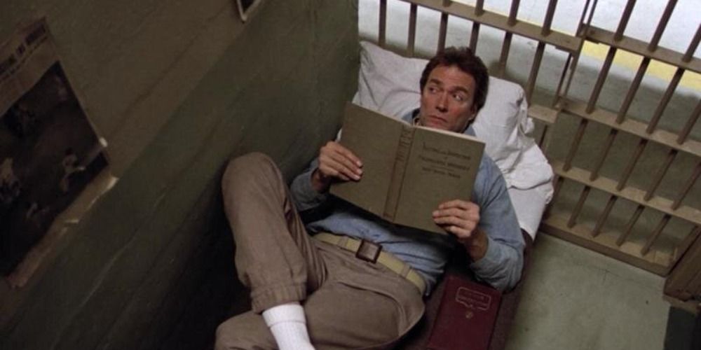 a man lying on a bed in a prison cell in Escape From Alcatraz, reading a large book