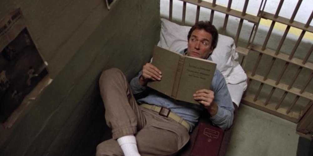 a man lying on a bed in a prison cell in Escape From Alcatraz, reading a large book