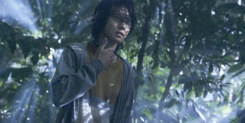 Arisu holding his neck as he stands in front of trees in Alice in Borderland.