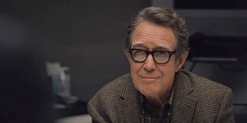 Sandy Furness from Succession wearing dark-rimmed glasses and a checkered sport coat, smiling.