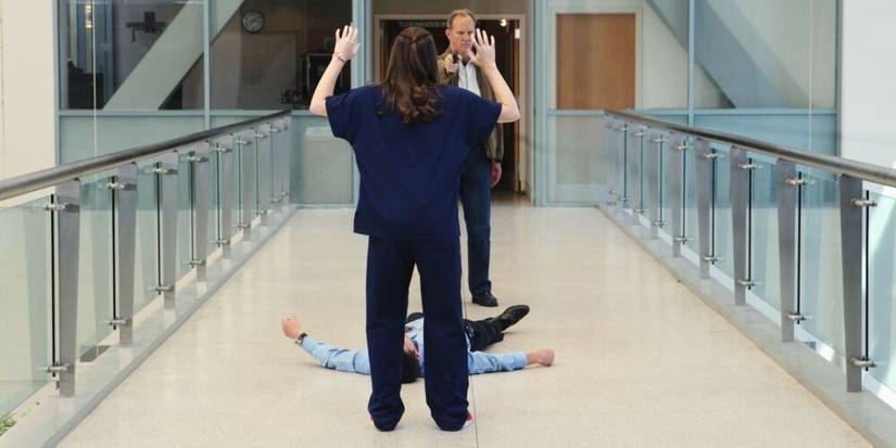 April Kepner with her back to the camera and hands up, in front of Derek Shepherd lying injured on the ground, and Gary Clark aiming a gun at April on Grey's Anatomy