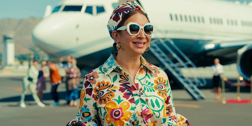 Maya Rudolph sorri usando uma camisa estampada e óculos escuros, em frente a um avião na série Loot.