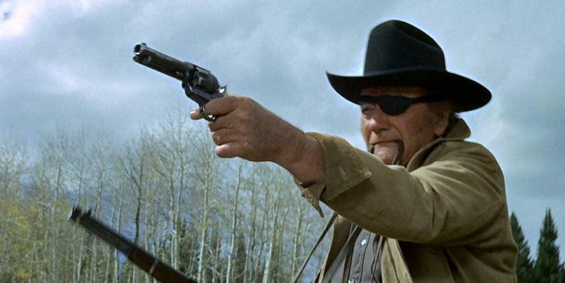 Imagem de Rooster Cogburn de John Wayne em True Grit (1969). A foto mostra Rooster empunhando um revólver e um rifle de repetição a cavalo, disparando para a esquerda da câmera.
