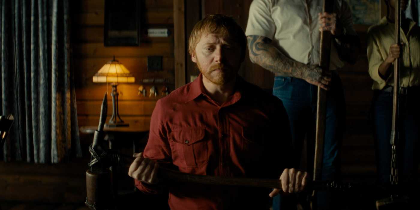 Rupert Grint kneeling with a weapon and looking sad in Knock at the Cabin