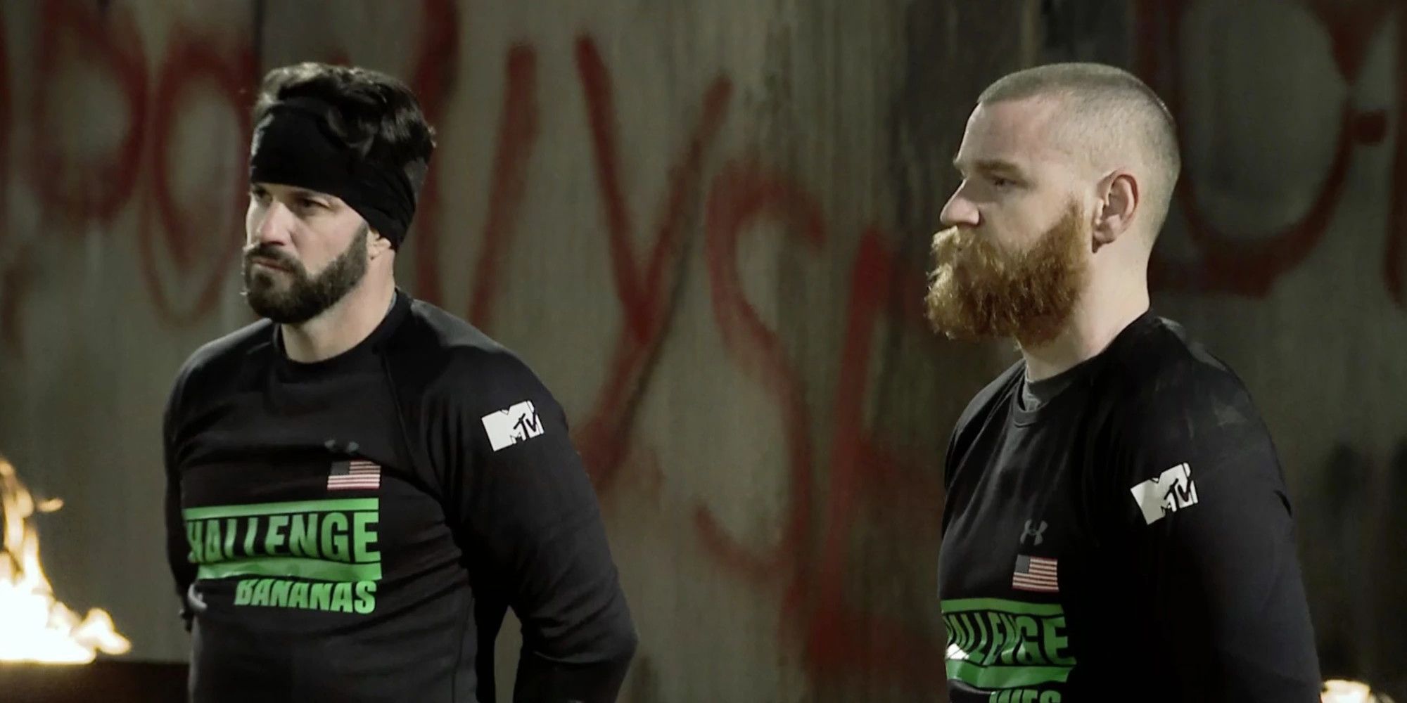 Bananas & Wes on The Challenge. They are standing beside each other and looking off camera, both wearing black long sleeve shirts with The Challenge logo written in green. They are in front of a wall with red lettering on it. 