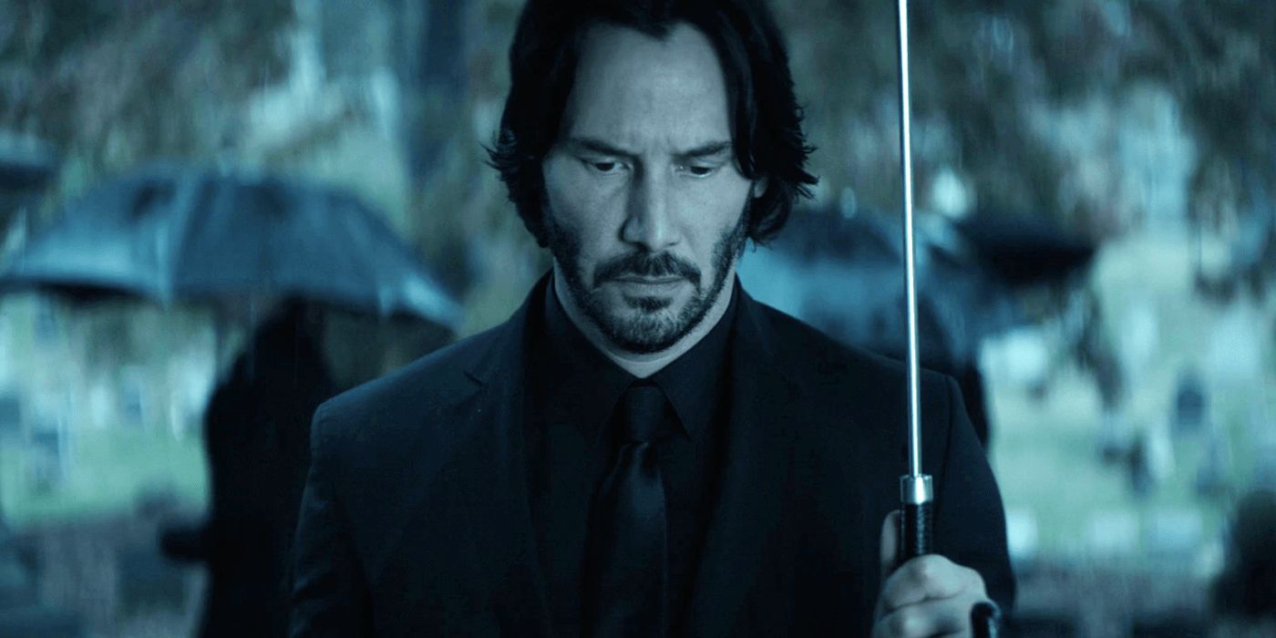 Screencap of Keanu Reeves as John Wick attending the funeral of his wife Helen in John Wick (2014). John is wearing a black suit and holding an umbrella as he looks down at Helen's grave. A few onlookers can be seen in the background, with rain also pouring.