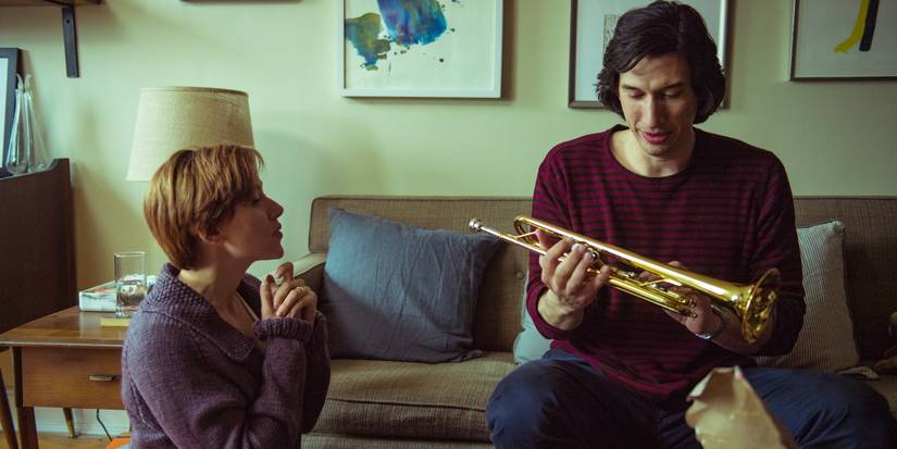 Adam Driver holds a trumpet and sits on a couch while Scarlett Johansson sits on the floor and watches him in a scene from Marriage Story