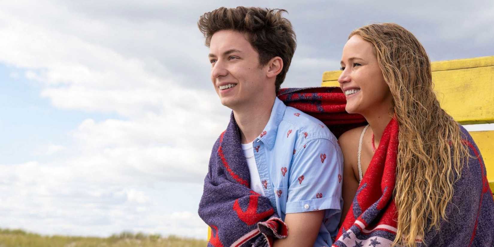 Jennifer Lawrence as Maddie and Andrew Barth Feldman as Percy have a towel around them while they sit at the beach in No Hard Feelings
