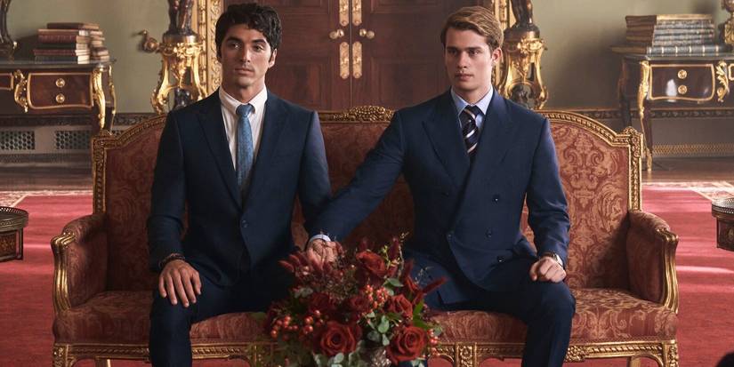 Alex (Taylor Zakhar Perez) and Prince Henry (Nicholas Galitzine) hold hands while sitting on a couch in Red White and Royal Blue.