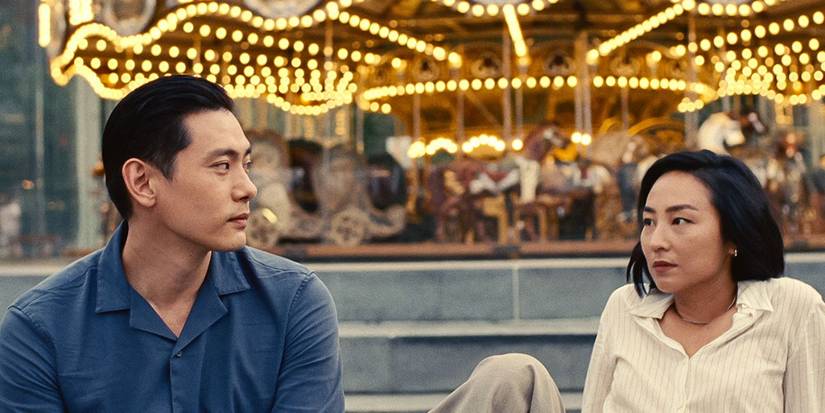 Nora (Greta Lee) e Hae (Teo Yoo) sentados em frente a um carrossel olhando um para o outro em Vidas Passadas