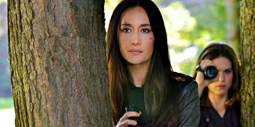 Maggie Q como Nikita atrás de uma árvore com outro agente usando uma câmera em Nikita