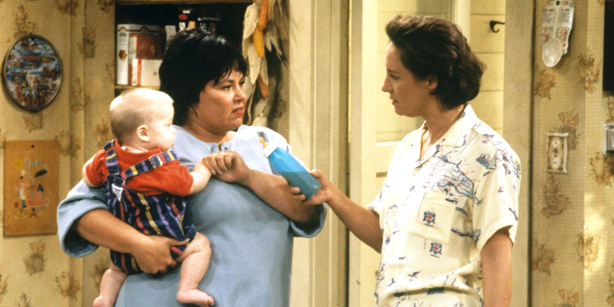 Roseanne (Roseanne Barr) with baby Jerry Garcia talking to Jackie (Laurie Metcalf) in the kitchen in Roseanne.