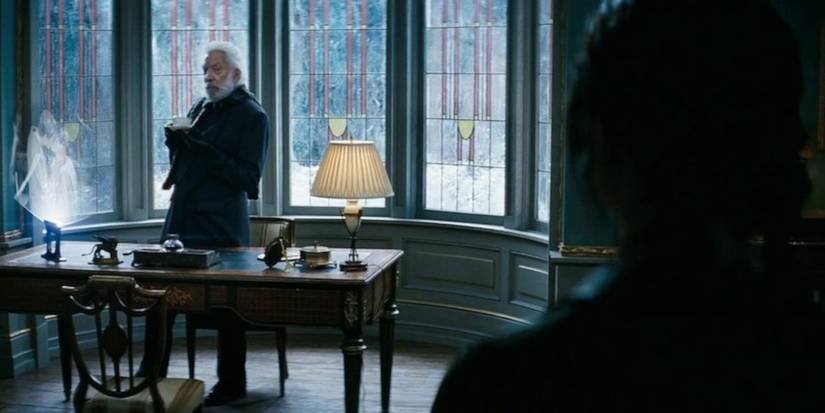 Donald Sutherland como Presidente Snow em pé atrás de uma mesa e olhando para Katniss nos Jogos Vorazes: Catching Fire