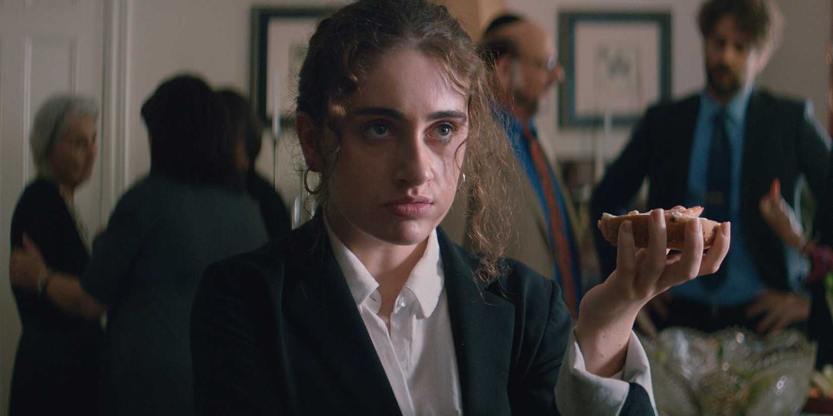 Danielle (Rachel Sennott) holding up a bagel at a shiva in Shiva Baby