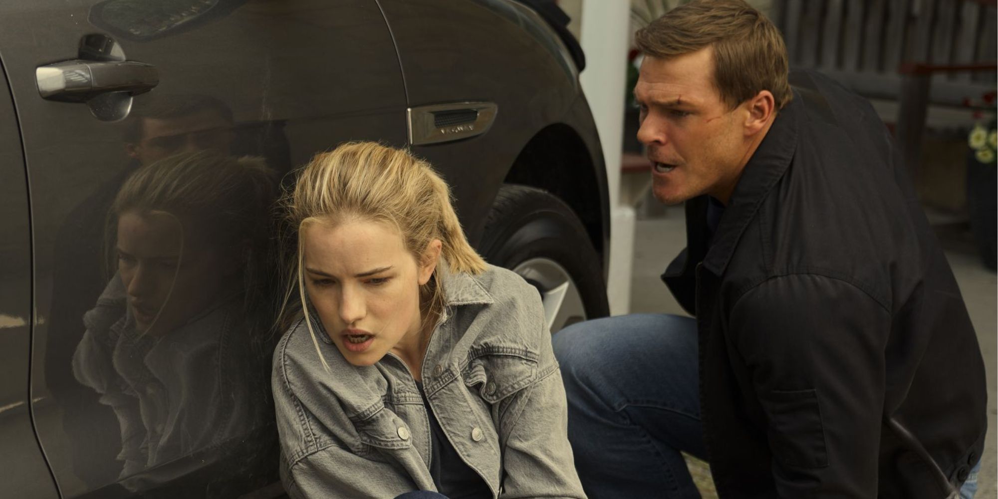 Willa Fitzgerald as Roscoe Conklin hiding behind a car with Alan Ritchson's Jack Reacher.