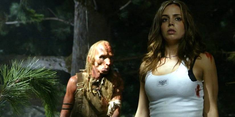 Eliza Dushku parecendo nervosa na floresta com um canibal atrás dela em O Massacre da Estrada 2003