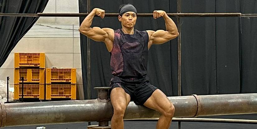 Amotti from Physical 100 flexes both arms wearing a black headband, black shorts, and black tank top.
