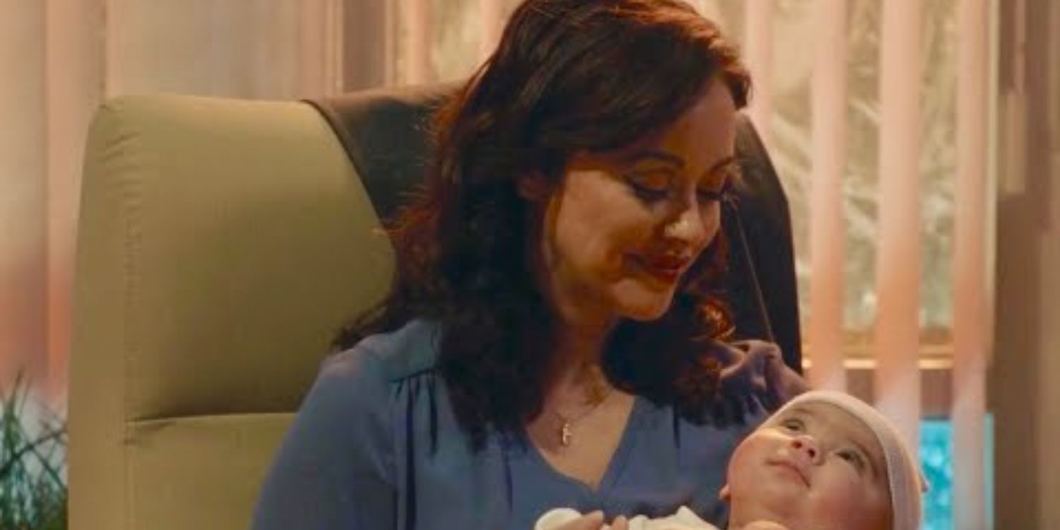 Blue Bloods Baez holds her baby. She is sitting in a green chair, wearing a blue shirt and looking down at the infant. There are open blinds behind her.