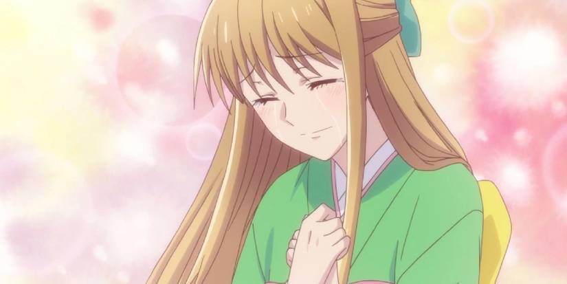 Fruits Basket's Ritsu cries as she clutches her chest with a pink and yellow background.
