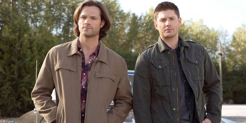 Sam (Jared Padalecki) and Dean Winchester (Jensen Ackles) leaning against their car and looking outward in Supernatural season 15.