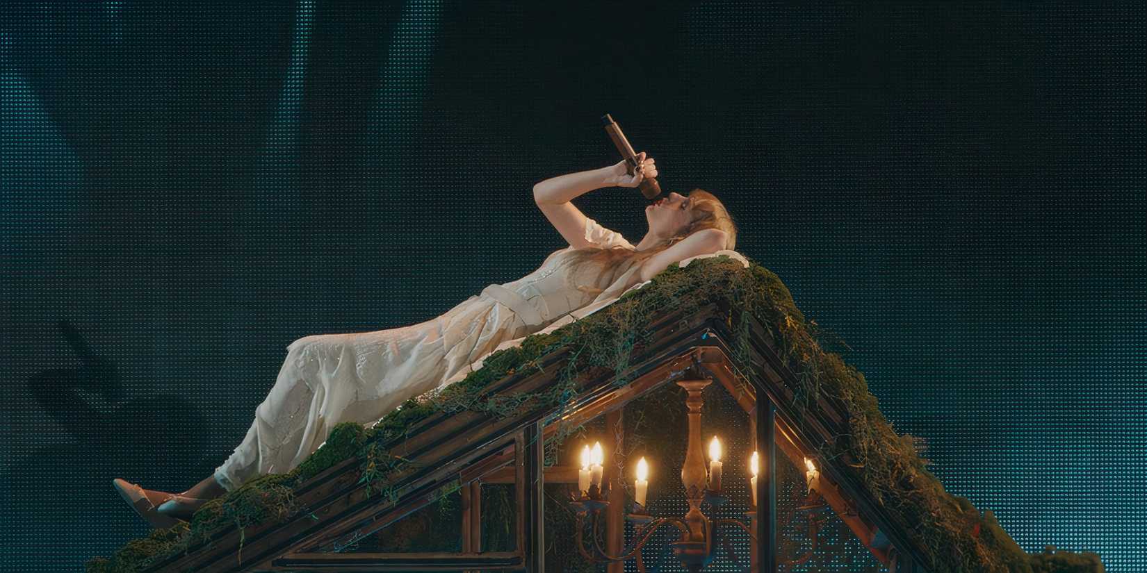 Taylor Swift lies on the mossy roof of the folklore cabin while singing "the 1" in The Eras Tour movie.