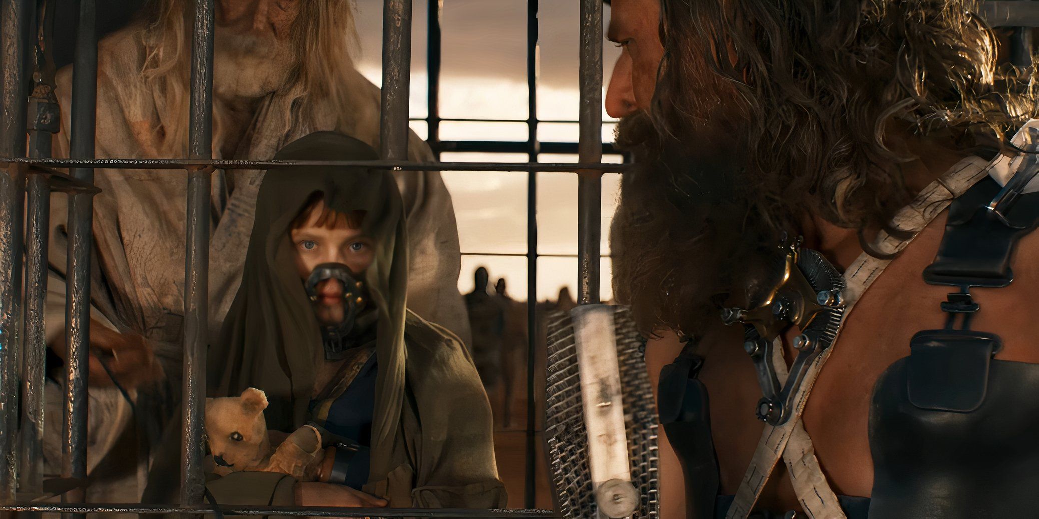 Young Furiosa holds a teddy bear while wearing a muzzle in a cage while Dementus (Chris Hemsworth) holds a microphone in Furiosa: A Mad Max Saga