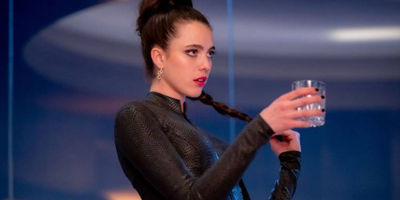 Сью (Мэгги Кэлли) держит свою длинную косу в одной руке и бокал в другой, одетая в черное боди с рептилиевой текстурой.