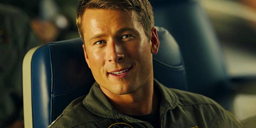 Glen Powell as Hangman sitting in a flight school chair smiling and looking up at someone in Top Gun Maverick