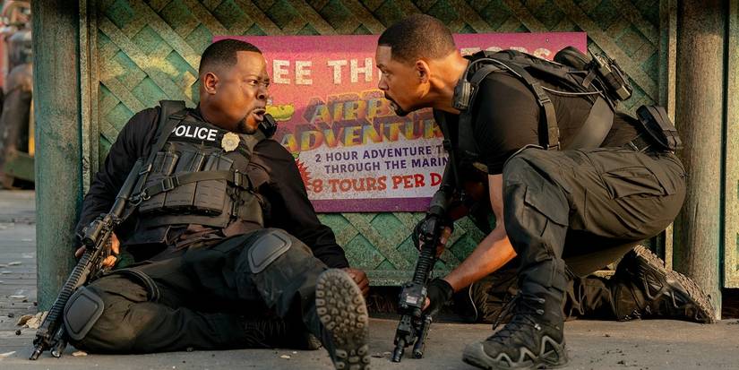 Marcus, sitting on the ground while leaning against a wall, yells at Mike, who crouches beside him holding a gun in Bad Boys Ride or Die