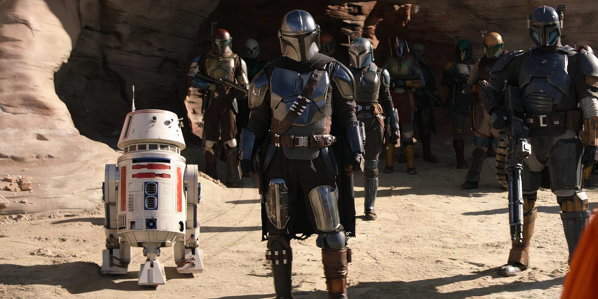 Din Djarin stands on the beach alongside R5-D4 and other Mandalorians