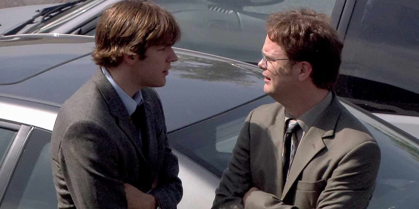 Jim (John Krasinski) and Dwight (Rainn Wilson) leaning against a car and talking in the parking lot in The Office season 1 episode "The Alliance."