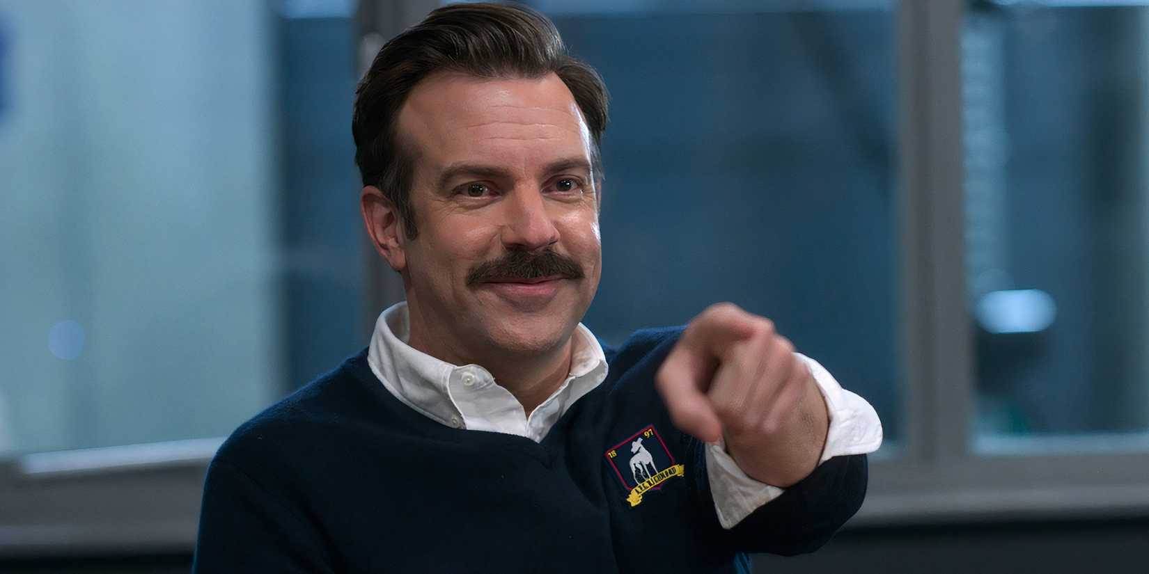 Jason Sudeikis como Ted Lasso sorrindo e apontando para algo fora de quadro em Ted Lasso.