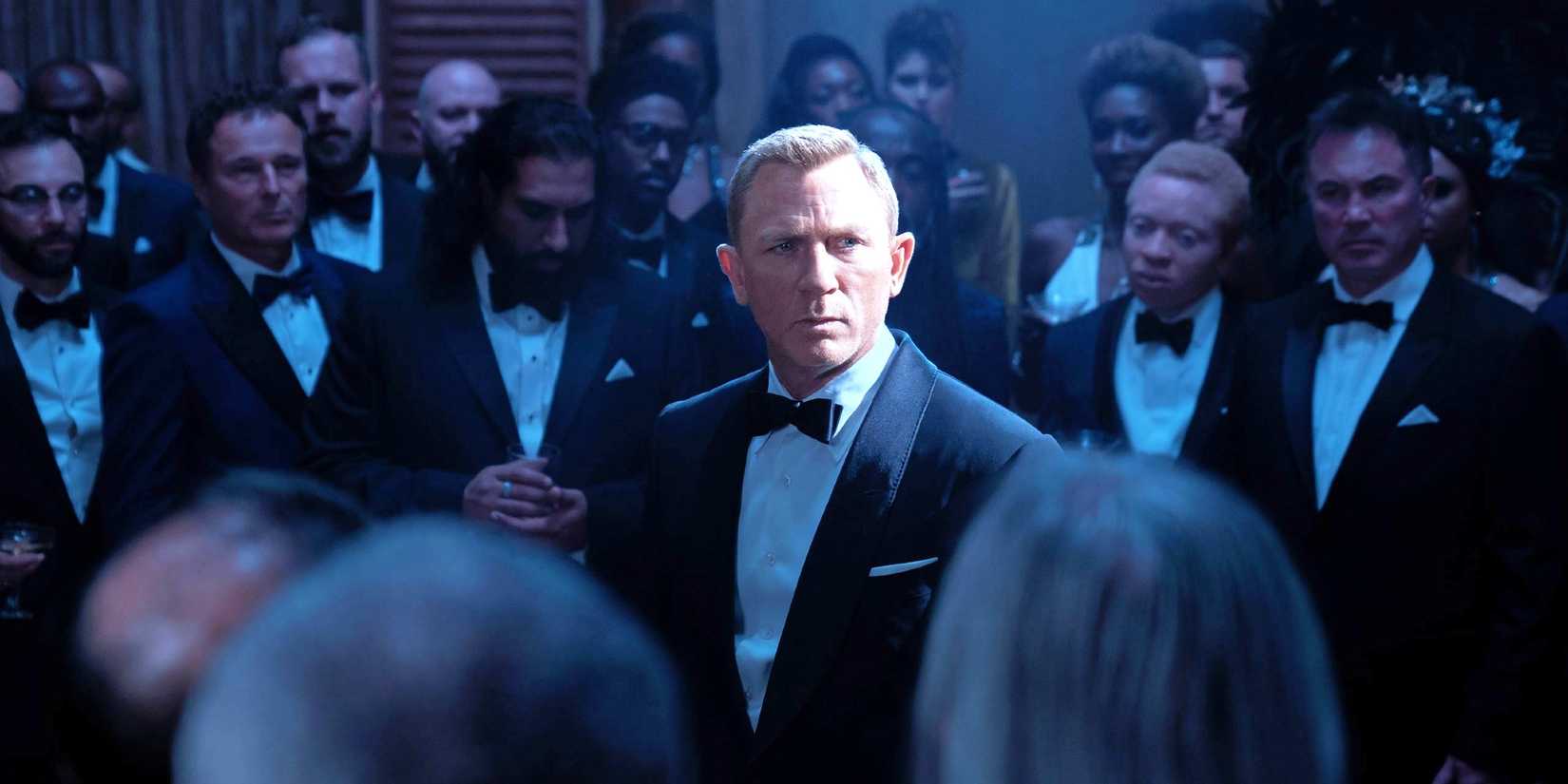 Daniel Craig as James Bond standing among a crowd in No Time to Die