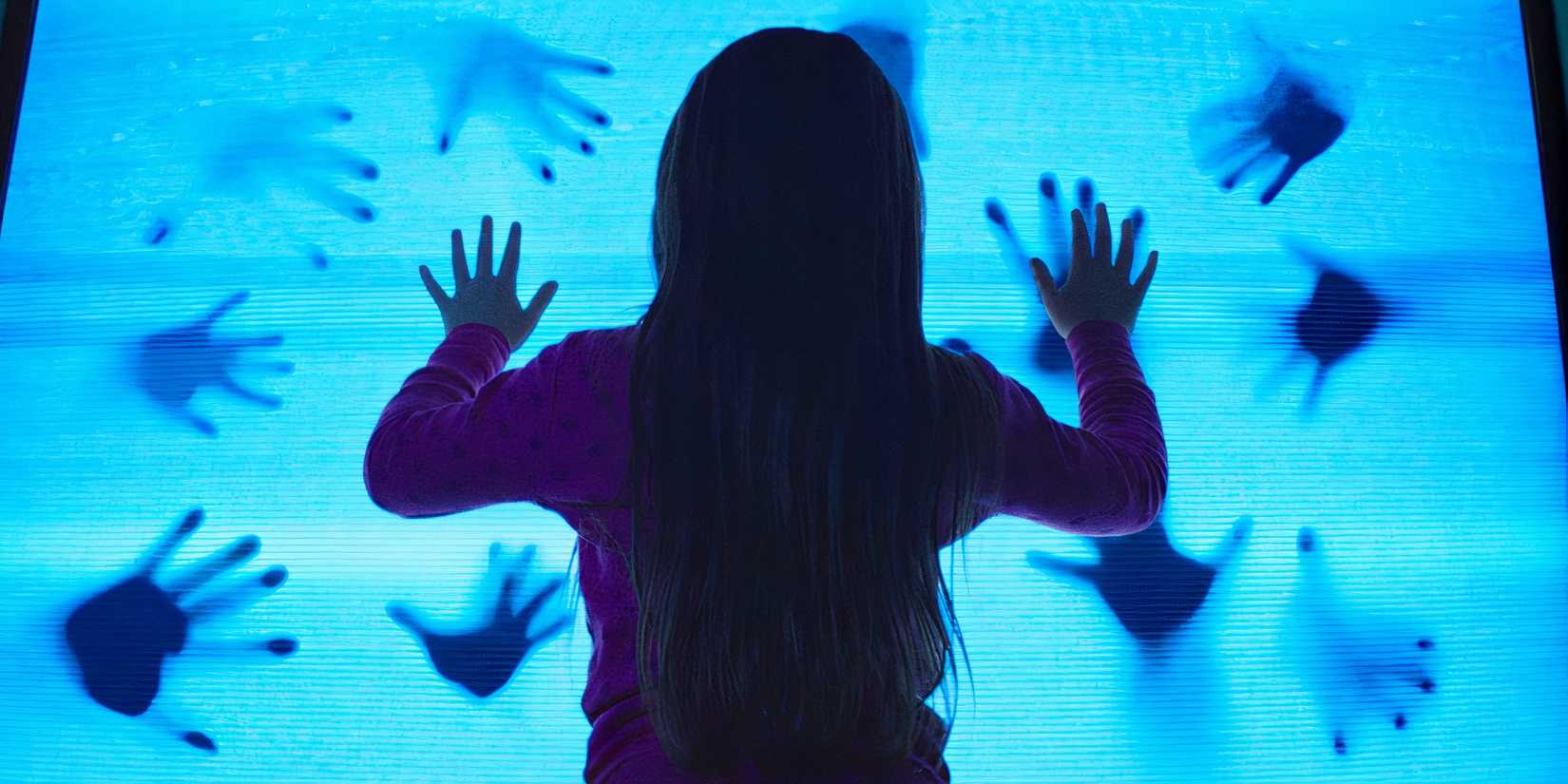 Atriz Kennedi Clements no remake de Poltergeist de 2015, com as mãos contra a tela da TV.