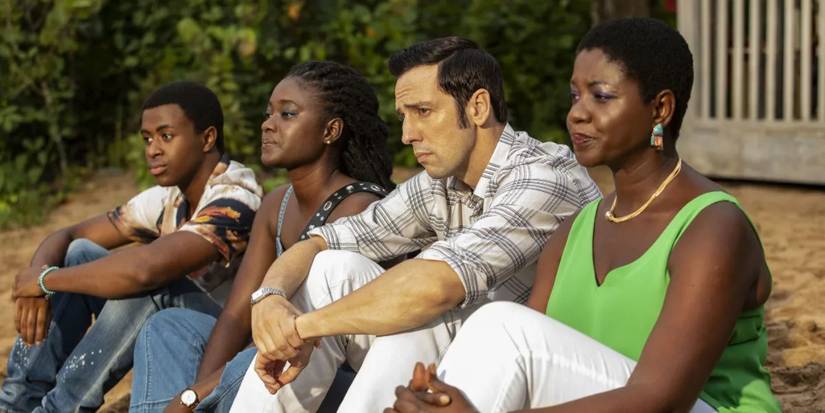 the cast of death in paradise sitting on the beach and looking upset
