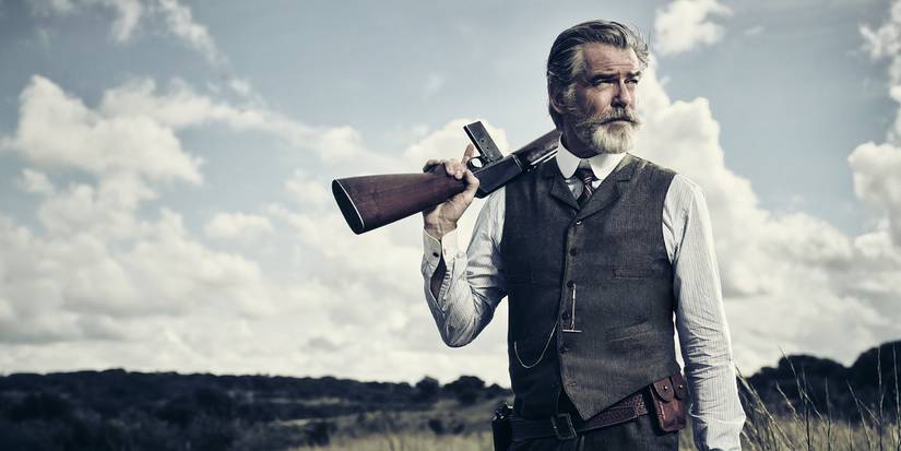 The Son, El (Pierce Brosnan) in a suit waistcoat with a gun slung over his shoulder 