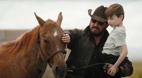 Yellowstone Rip holding a little boy while petting a horse with his other hand