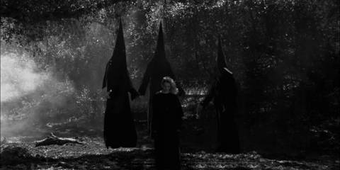 A coven of three witches in pointed black hoods gathers around a girl in the woods.