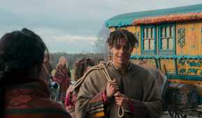 Aram stands in front of a colorful Tuatha'an wagon in The Wheel of Time