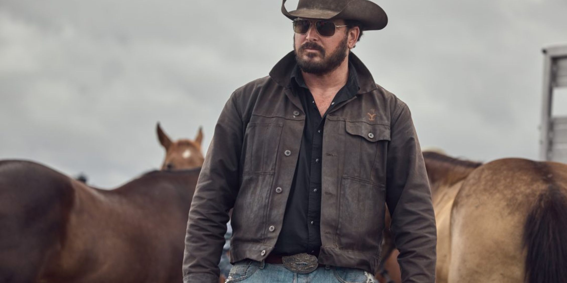 Yellowstone Rip wearing sunglasses and cowboy hat looking out at the cowboys while standing with horses