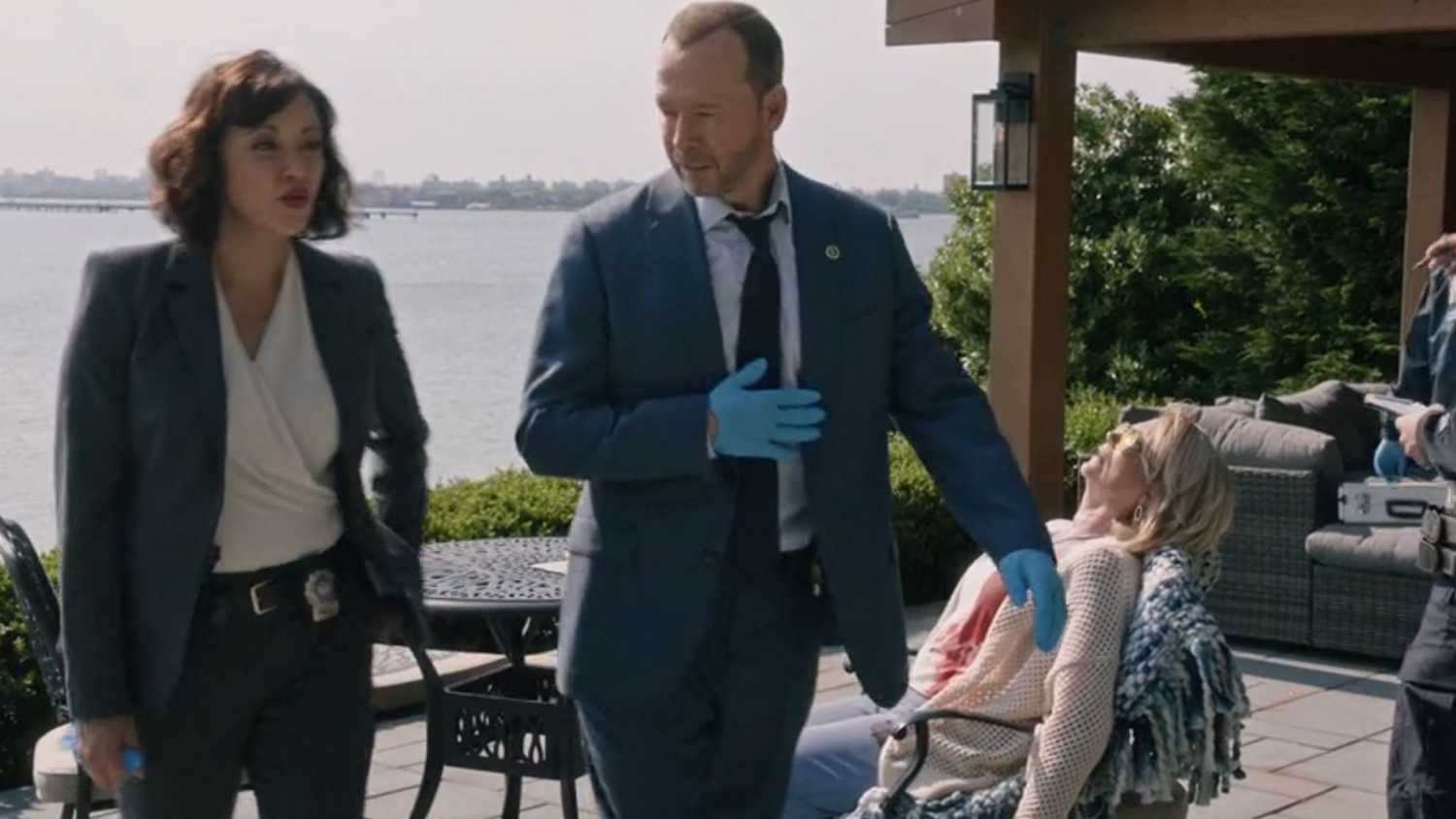Blue Bloods Danny And Baez wearing blue gloves while a dead body sits behind them in a deck chair