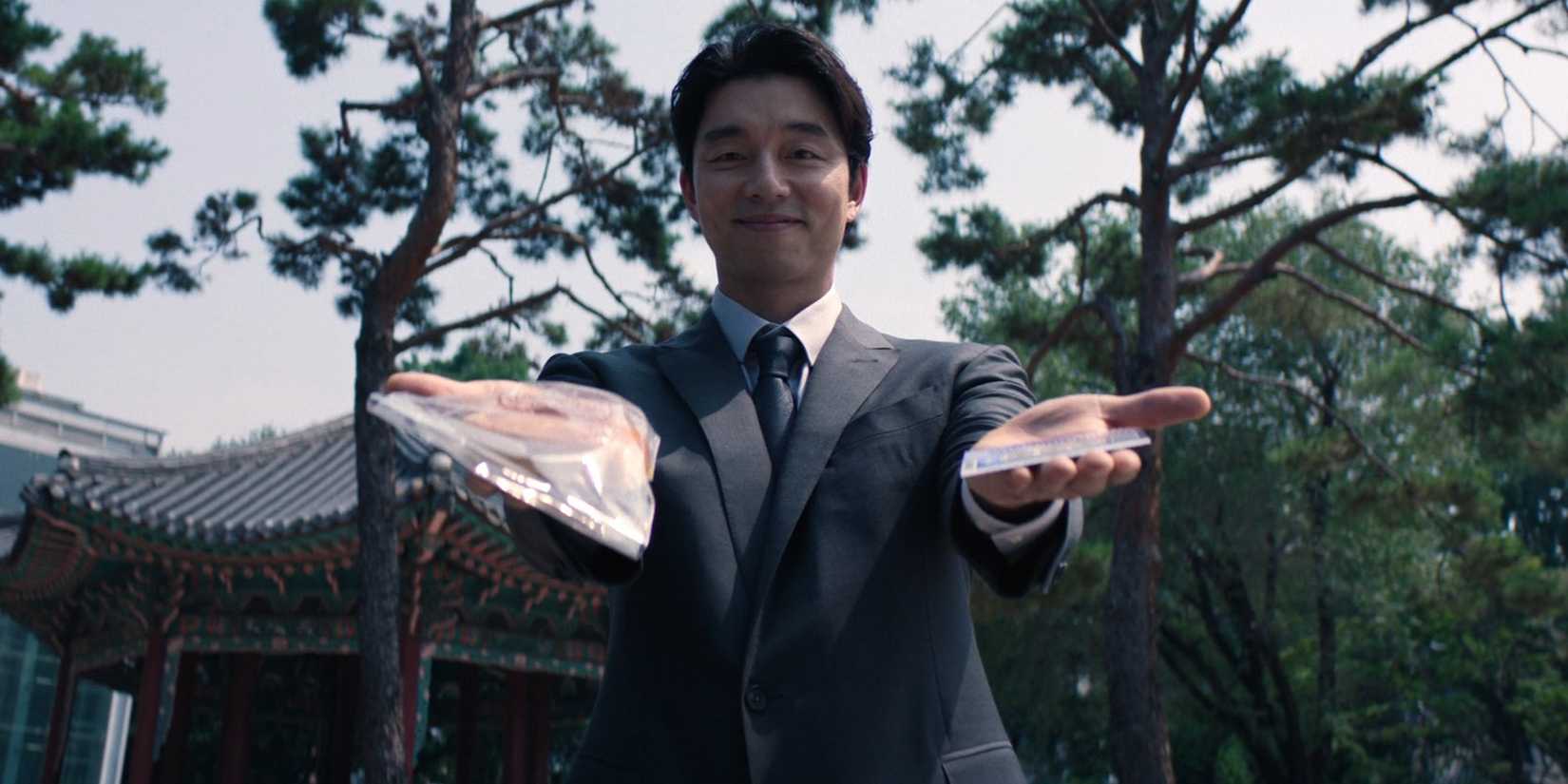 The Salesman (Gong Yoo) holds out bread in one hand and a lottery ticket in the other in Squid Game season 2, episode 1 "Bread and Lottery"