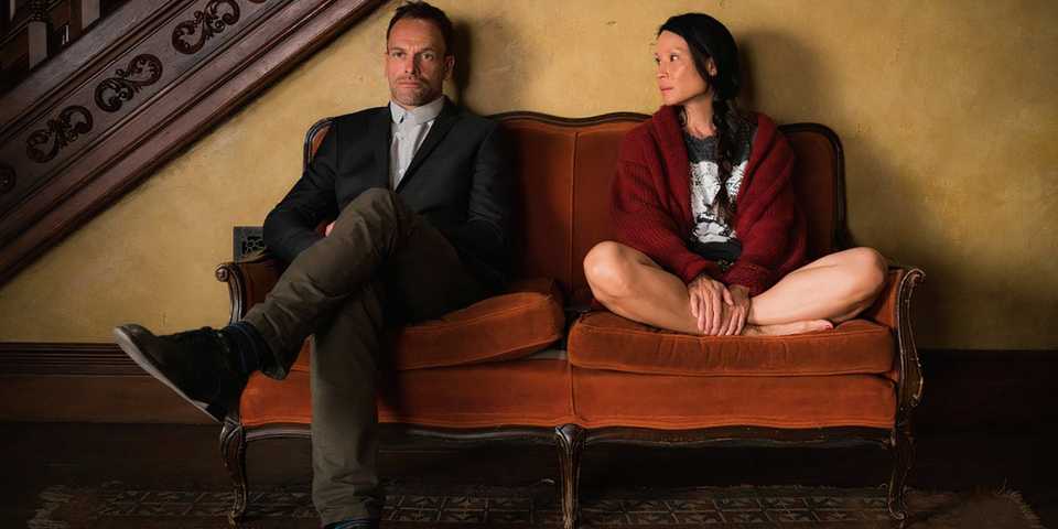Jonny Lee Miller's Sherlock and Lucy Liu's Joan sitting on a couch with each other in Elementary.
