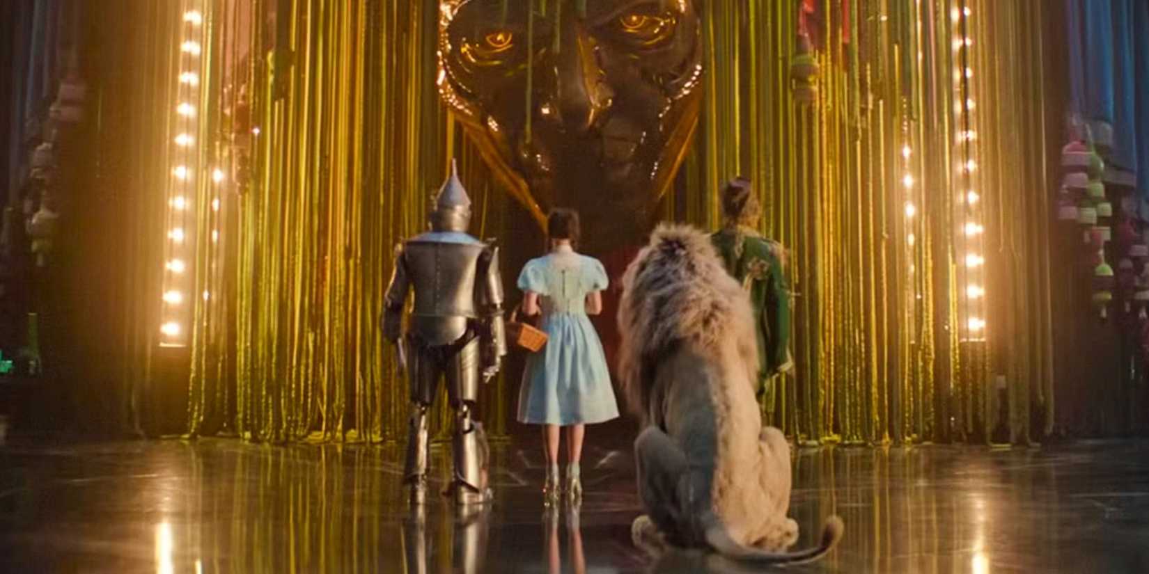 Dorothy, Tin Man, Scarecrow, and Cowardly Lion shown from behind as they face the Wizard's metal head in Wicked