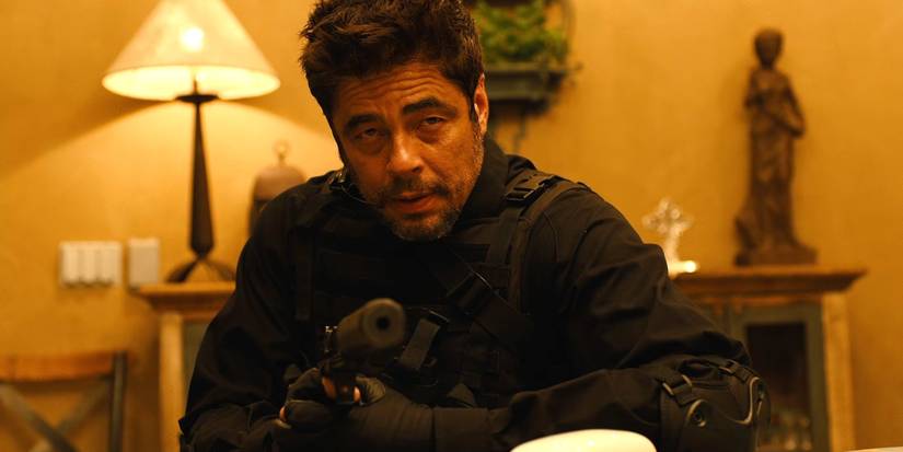 Benicio del Toro como Alejandro segurando uma pistola silenciada e vestindo um traje furtivo em Sicario