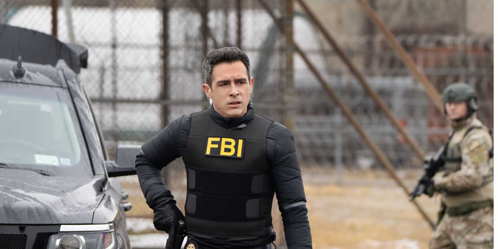 Scola do FBI usando seu colete do FBI e segurando uma arma com homens em equipamento militar atrás dele
